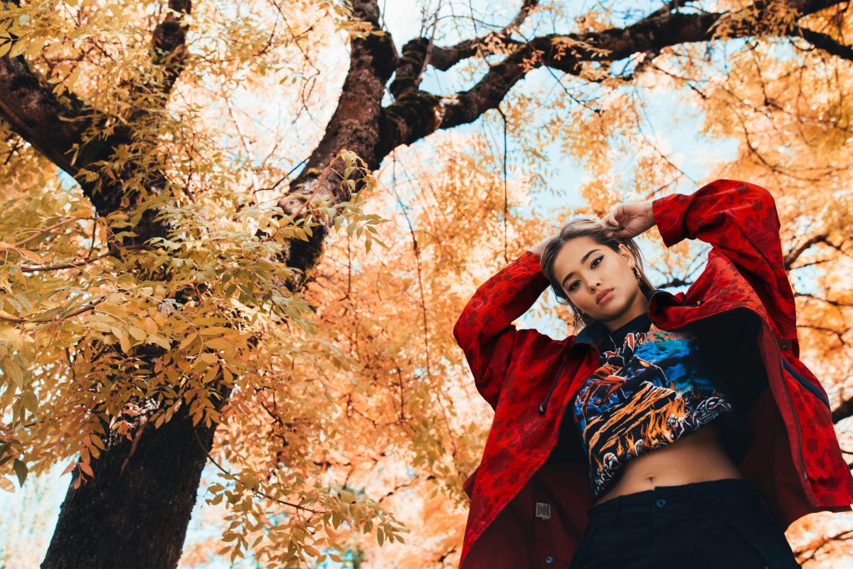 A stylish woman wearing a red jacket and graphic t-shirt poses confidently under a tree with golden autumn leaves, reflecting the vibrant fall fashion trends of 2024.